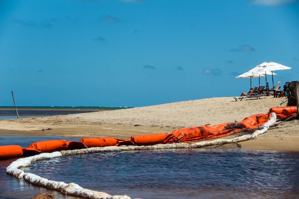 Uma turista toma sol ao lado da foz do rio Persinunga, que delimita a fronteira (na região litorânea) entre os estados de Pernambuco e Alagoas. A boia de contenção tenta evitar que o óleo se espalhe do mar em direção ao rio.