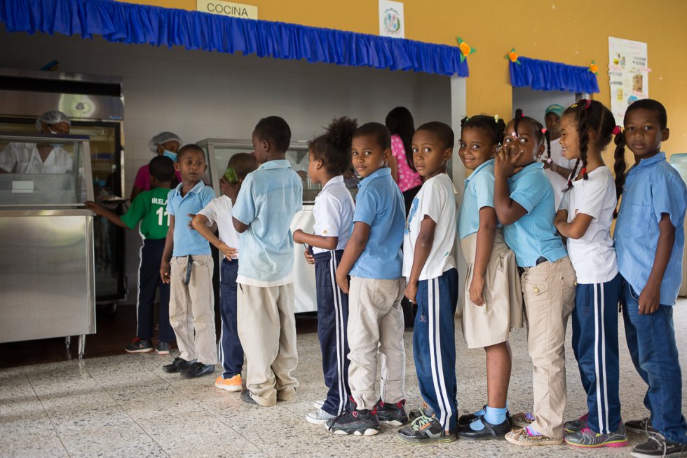 En la provincia de Monte Plata (República Dominicana), se desarrolla un programa piloto para garantizar la seguridad alimentaria que luego pretende ser escalado a todo el país. Se trata de crear un círculo virtuoso en el que la comida que se sirve en los colegios públicos se compre a cooperativas de pequeños agricultores locales, potenciando la producción y garantizando un mercado para estos últimos al tiempo que se asegura que los niños reciban una nutrición adecuada y sus familias un desahogo económico. En la imagen, un grupo de alumnos hace fila para el almuerzo.