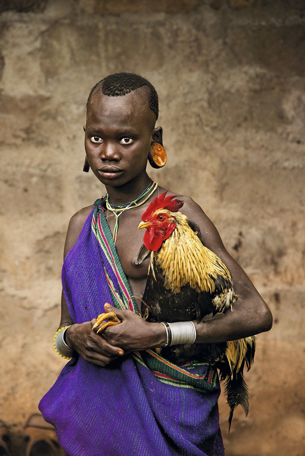 Uma garota da tribo Kara do Vale do Omo (Etiópia) segura um galo. As garras do animal aparecem, pacíficas, nas mãos da criança. Ano 2013.