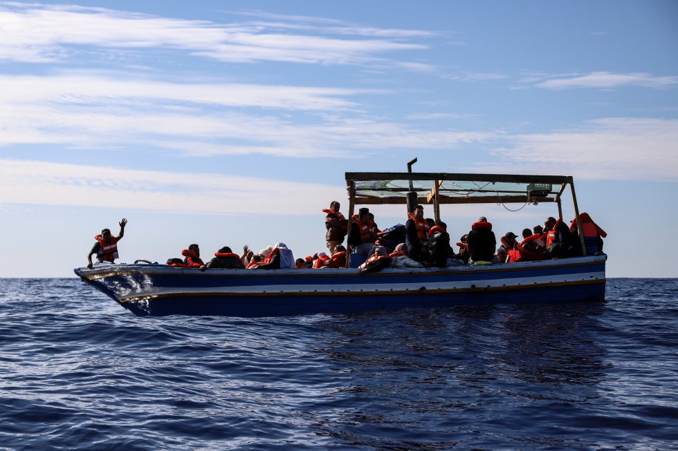 Un migrante hace señas con la mano mientras otros permanecen sentados en una precaria barca de madera antes de ser rescatados por un equipo de 'Sea Watch 3' en la zona SAR de Libia, a unas 30 millas de la costa, el 17 de octubre de 2021. En las 48 horas siguientes a ese día, el Sea Watch 3 rescató a 412 migrantes de siete embarcaciones en apuros.