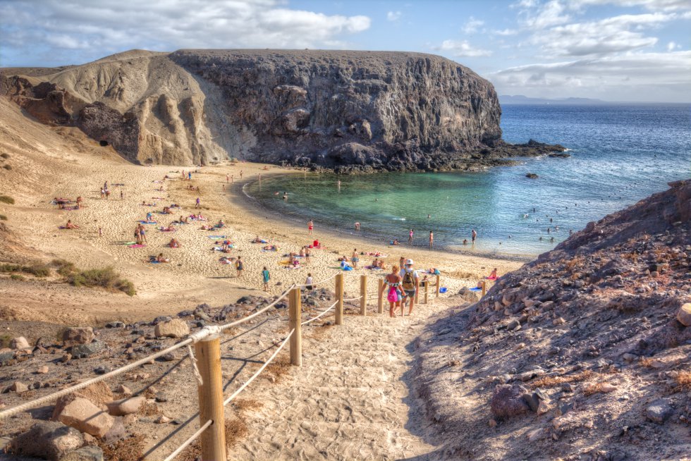 El sureste de Lanzarote se ve dorado en su arena, se siente frío por un Atlántico de tonos azules verdosos, sabe a salitre, suena a viento alisio y huele a arena quemada por el sol africano. Con Fuerteventura y el islote de Lobos al fondo, la caleta de Papagayo, la más fotogénica y aplacerada, la que da nombre al resto de playas del monumento natural de los Ajaches, sirvió hasta mediados del siglo pasado como fondeadero. Los niños gustan de su media luna bien trazada; otros practican snorkel en sus laterales. Pero todo se revela secundario frente a la panorámica que se divisa desde la terraza natural que otorga el acantilado, junto a El Chiringuito. El placer visual encuentra durante el crepúsculo su máxima expresión. Acceso: desde el enclave de Playa Blanca sale un ramal primero de asfalto y luego de tierra. Peaje medioambiental (3 euros), solo a coches, de 9.00 a 16.00 (hasta las 17.30 a partir de junio). Alquilan bicicletas en Papagayo Bike (papagayobike.com). La bajada ha sido acondicionada con escalones y postes de madera ensogados.