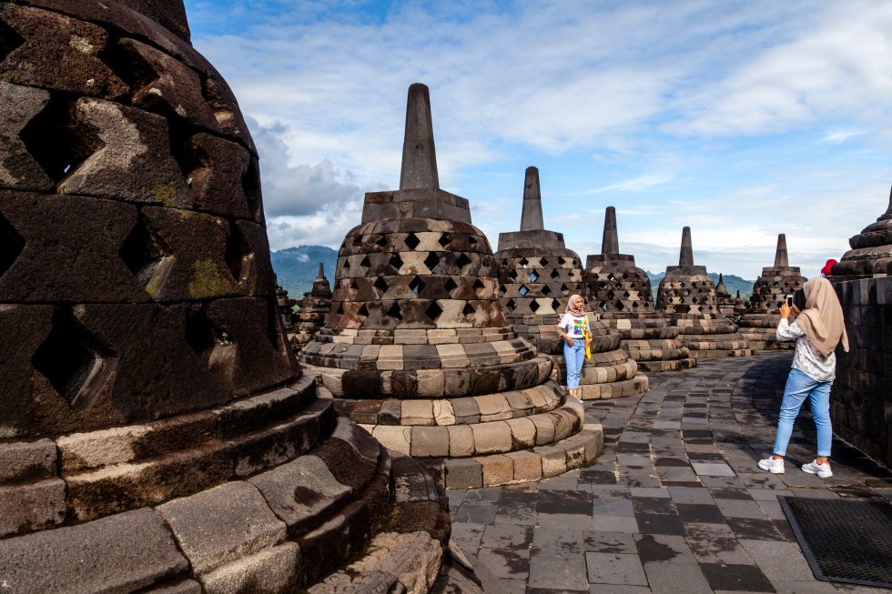 Junto con Angkor Wat en Camboya y Bagan en Myanmar, Borobudur hace que el resto de los espectaculares yacimientos del sudeste asiático casi parezcan insignificantes. Borobudur, que se alza por encima de los arrozales como una montaña, es un colosal monumento que resulta tan enigmático y hermoso como debió de ser hace más de 1.200 años. Es el templo budista más grande del mundo, construido con más de dos millones de bloques de piedra. Como un mandala en tres dimensiones, rematado por pisos de estupas perforadas, este complejo de roca de lava gris es el reflejo de los volcanes que se alzan a su alrededor, aunque al ir subiendo por cada piso lo que atrapa la mirada no son las vistas, sino los bajorrelieves de sus paredes. Talladas hasta el más mínimo detalle pueden verse escenas de la vida de Buda e ilustraciones vívidas de la ley del karma. En lo alto, la cúpula principal se alza como una flor de loto petrificada rodeada de estupas, algunas de las cuales han perdido sus remates y revelan estatuas de Buda en sus centros. Es una geometría ritual a escala monumental; cada medida y cada ángulo transmiten un poderoso mensaje.